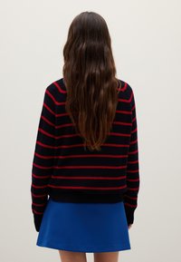 Maglione blu marina con strisce orizzontali rosse, texture lavorata a maglia, maniche lunghe, abbinato a una gonna blu. La vista dal retro mostra i capelli che cadono.