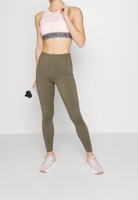 Reggiseno sportivo rosa chiaro con parte superiore in rete e banda con logo nera; leggings verde oliva con tasche laterali e dettaglio del logo; sneakers grigie.