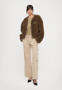 Frau in brauner Fleecejacke, beigen Cargo-Hosen und schwarzen High Heels, die mit den Händen in den Taschen der Jacke vor einem weißen Hintergrund steht.