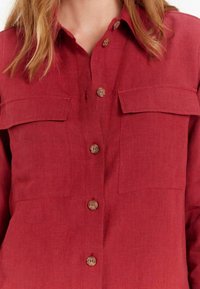 Chemise en lin rouge avec deux poches poitrine, fermeture à boutons et boutons contrastants marron clair. Présente un col classique et un tissu texturé.