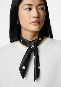 Zwarte zijden sjaal met witte polkadots, om de nek gebonden, met een gladde textuur en een subtiele glans. Draagt over een wit bovenstuk.