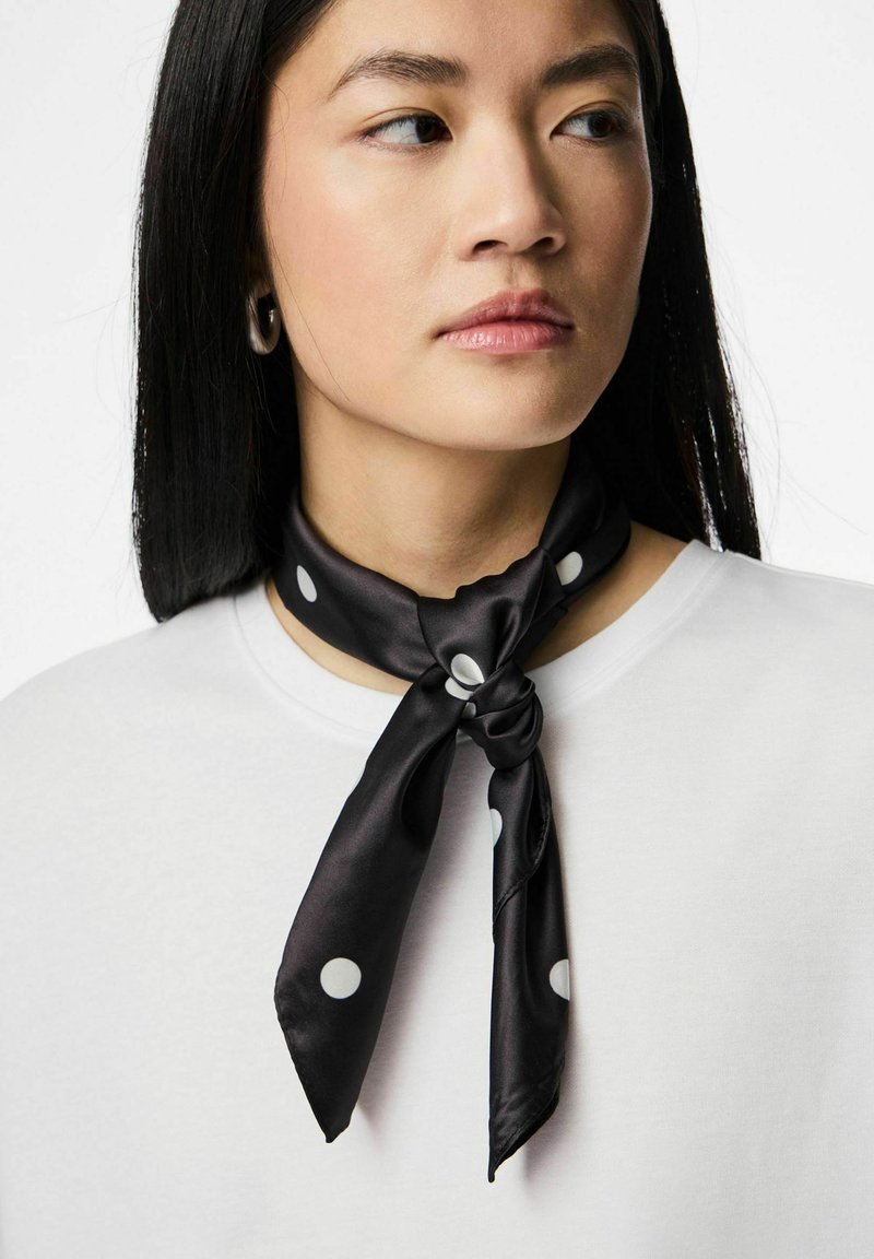 Zwarte zijden sjaal met witte polkadots, om de nek gebonden, met een gladde textuur en een subtiele glans. Draagt over een wit bovenstuk.