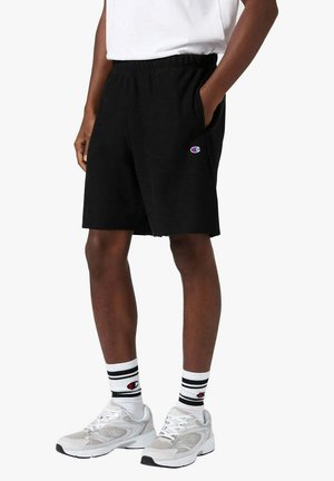 Persona che indossa pantaloncini neri Champion con le mani nelle tasche, calzini bianchi Champion con strisce nere e scarpe da ginnastica bianche.