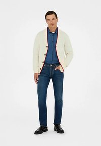 Krém színű kötött kardigán piros szegéllyel, kék farmeringgel, sötétkék farmerrel és fekete cipővel párosítva. A textúrákra és a színkontrasztra helyezve a hangsúlyt.