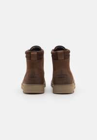Botine din piele maro, cu un vârf moale care se pliază, finisaj texturat și talpă din cauciuc. Prezintă un patch cu logo la spate.