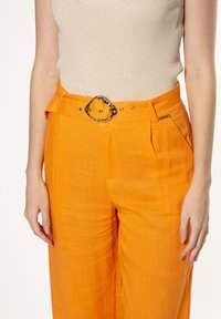 Oranje linnen broek met een wijde pasvorm, voorzien van een zilveren ovale riemgesp en frontplooien. De stof heeft een gestructureerde uitstraling.