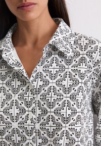 Primer plano de una persona que lleva una camisa blanca de botones con un patrón de cuadrícula floral negra y el collar abierto.
