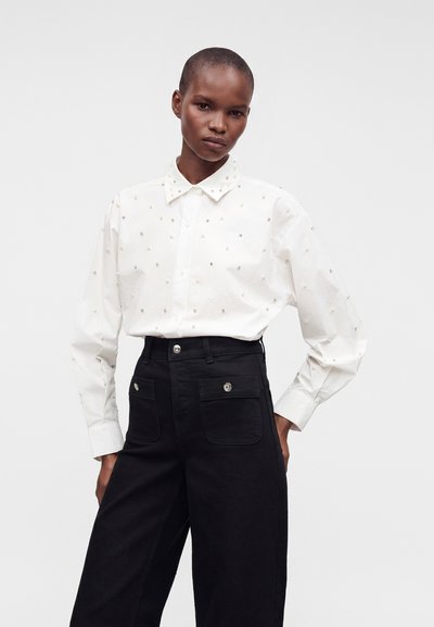 Femme portant une chemise blanche à boutons avec de petites décorations en perles et un pantalon noir taille haute avec des poches avant, debout devant un fond uni.