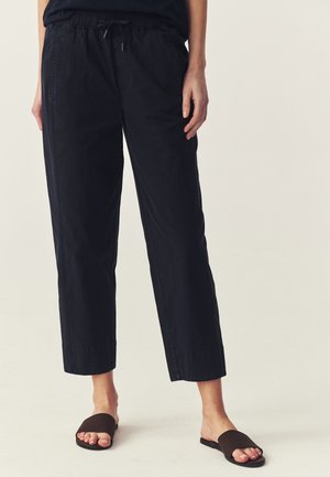Persona que lleva pantalones negros recortados con cintura elástica y sandalias negras abiertas, de pie sobre fondo claro.