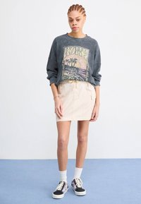 Grijze oversized sweatshirt met palmbomenprint, crèmekleurige minirok, witte sokken en zwarte sneakers met paarse accenten. Effen achtergrond.