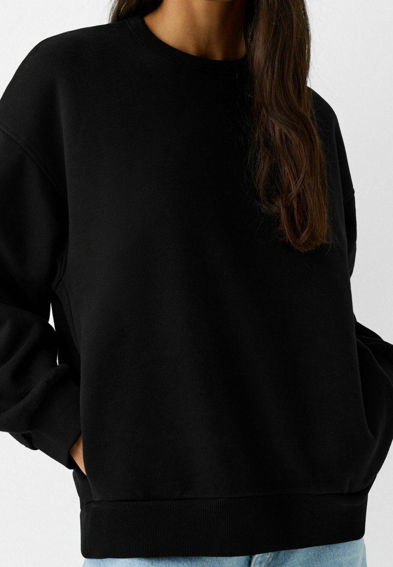 Personne portant un sweat-shirt noir à col rond, les mains dans les poches et de longs cheveux bruns tombant sur une épaule.