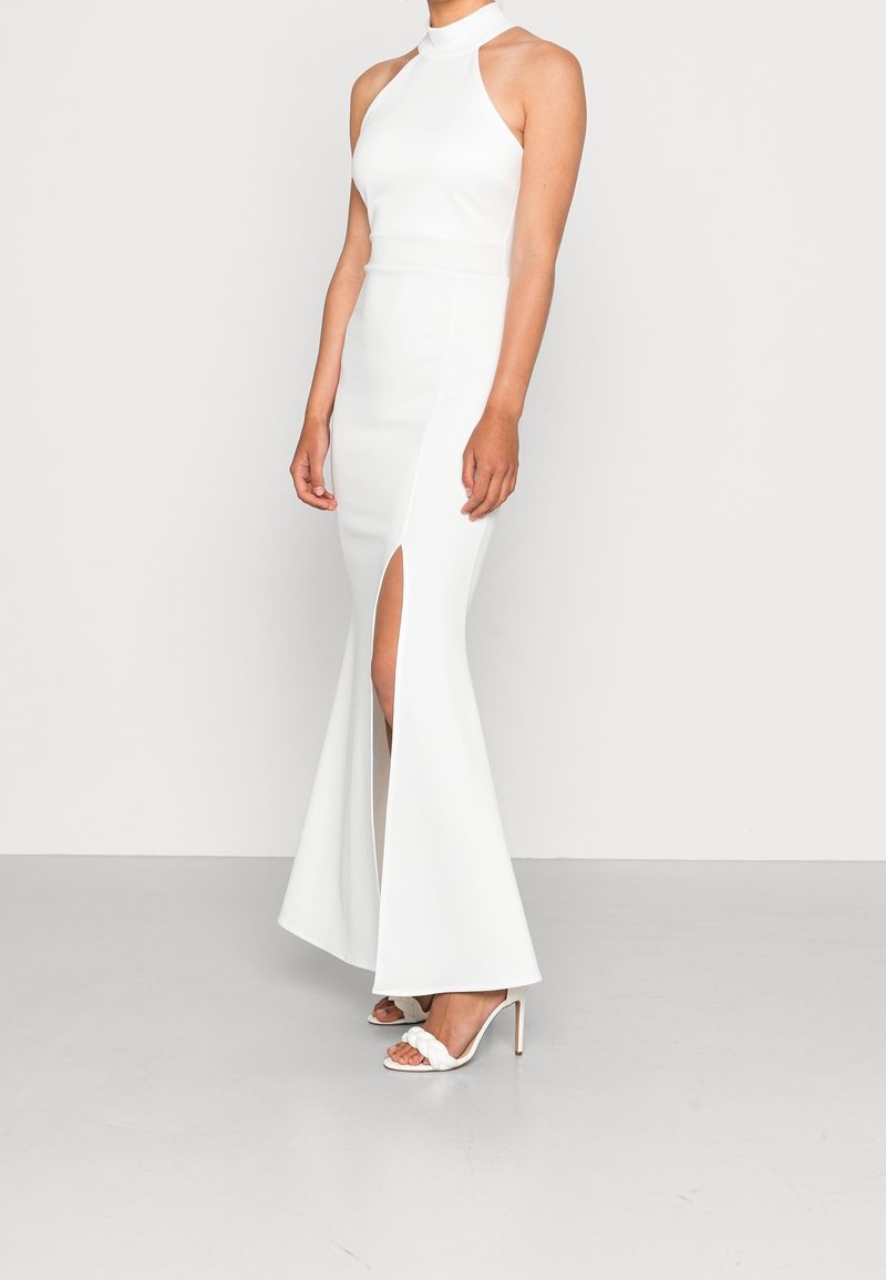 Robe blanche à halter avec fente latérale, silhouette ajustée et bas évasé. Associée à des sandales à talons hauts blanches. Texture lisse.