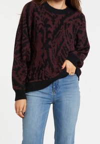 Suéter oversize burdeos y negro con un estampado, acabado texturizado, cuello redondo y puños acanalados, combinado con jeans de color azul claro.