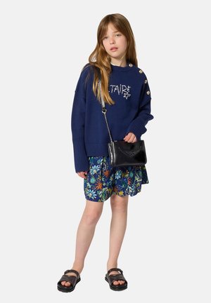 Jong meisje dat een marineblauwe trui met knopen draagt, een bloemenrok, een zwarte crossbodytas en zwarte sandalen, staand tegen een effen achtergrond.