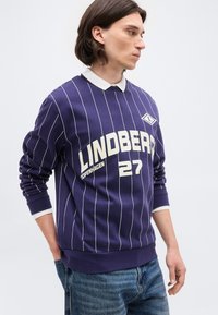 Άνδρας με καστανά μαλλιά φοράει μπλε σκούρο πουλόβερ με λεπτές ρίγες και το κείμενο «LINDBERG 27 COPENHAGEN», λευκό γιακά και μπλε τζιν, κοιτάζοντας προς τα δεξιά.