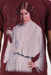 Maroon t-shirt featuring a graphic print of a character in a white robe with gathered waist, holding a blaster. Texture appears soft.