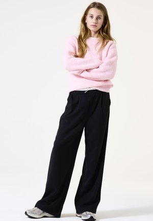 Un pull en tricot côtelé rose, un pantalon large noir avec taille élastique, et des baskets argentées. Une tenue simple et décontractée avec une silhouette relaxée.