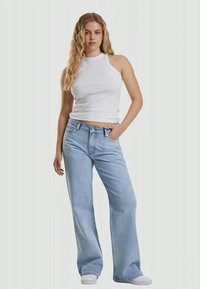 Joven mujer con cabello largo y rizado de pie, vistiendo una camiseta blanca sin mangas, pantalones vaqueros anchos de color azul claro y zapatillas blancas.