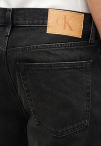 Εγγύς πλάνο πίσω τσέπης μαύρου τζιν με ταμπά δερμάτινο έμβλημα με το λογότυπο "cK" κοντά στη μέση.