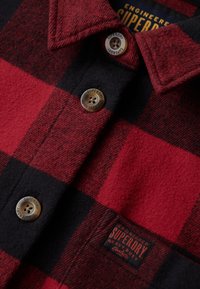 Camicia in flanella a quadri rossi e neri con design a bottoni, colletto arrotondato e bottoni metallici marchiati. Etichetta tessuta sull'orlo in un colore a contrasto.
