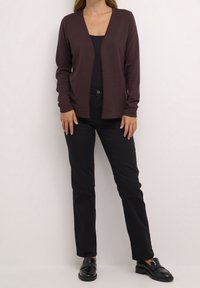 Cardigan marron avec un col en V profond, manches longues et une texture douce, associé à un pantalon noir ajusté et des mocassins noirs.