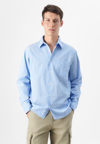 Un joven se muestra con confianza en una camisa de botones azul a rayas y pantalones beige, sonriendo suavemente frente a un fondo blanco.