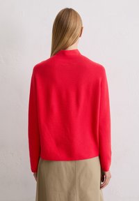 Roter gestrickter Pullover mit hohem Kragen und langen Ärmeln, mit lockerem Schnitt und glatter Textur, kombiniert mit einem beigen Rock.