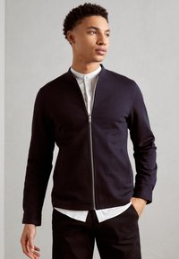 Mornarica zip-up sweatshirt iz mehkega blaga z zaobljenim ovratnikom, oprijet kroj in dolge rokave, oblečen čez belo srajco s kragnico.