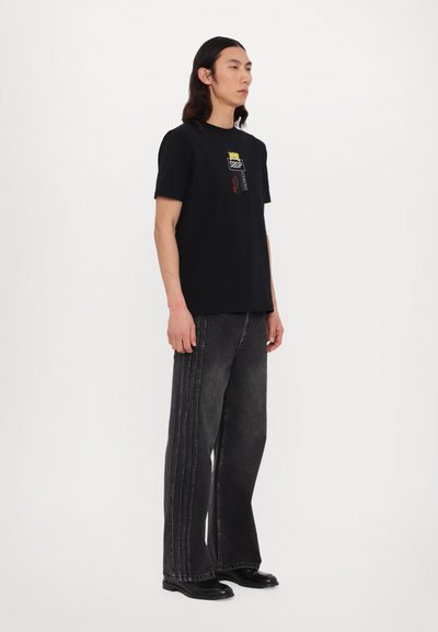 Sort bomulds T-shirt med korte ærmer, der har et grafisk design i gul og rød. Sammen med mørkegrå brede bukser og sorte sko.