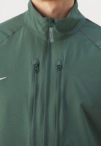 Vihreä takki korkealla kauluksella, jossa on kaksi etutaskua vetoketjulla, täysi vetoketju ja pieni valkoinen logo vasemmassa rinnassa. Sujuva pinta.