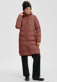 Vrouw in een koperen lange donsjas, zwarte broek, zwarte laarzen en een zwart hoofdband, staand met één hand bij haar kraag.