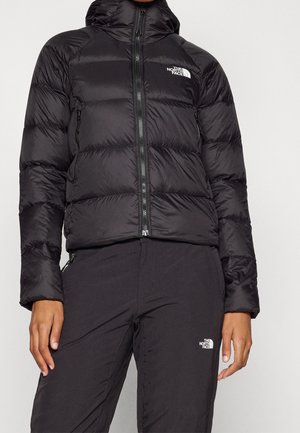 Persona vistiendo una chaqueta acolchada negra de The North Face y pantalones negros, ambos con logotipos visibles de la marca, sobre un fondo claro y liso.