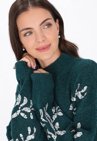 Türkisfarbener Strickpullover mit langen Ärmeln, der mit weißen Paillettenblumenmustern verziert ist. Der Stoff wirkt weich, hat gerippte Bündchen und einen hohen Ausschnitt.
