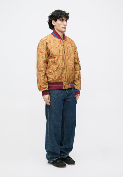 Zlatá bomber bunda s červenými akcentmi a vzorovaným dizajnom. Nosená s voľnými modrými rifľami a čiernymi plochými topánkami.