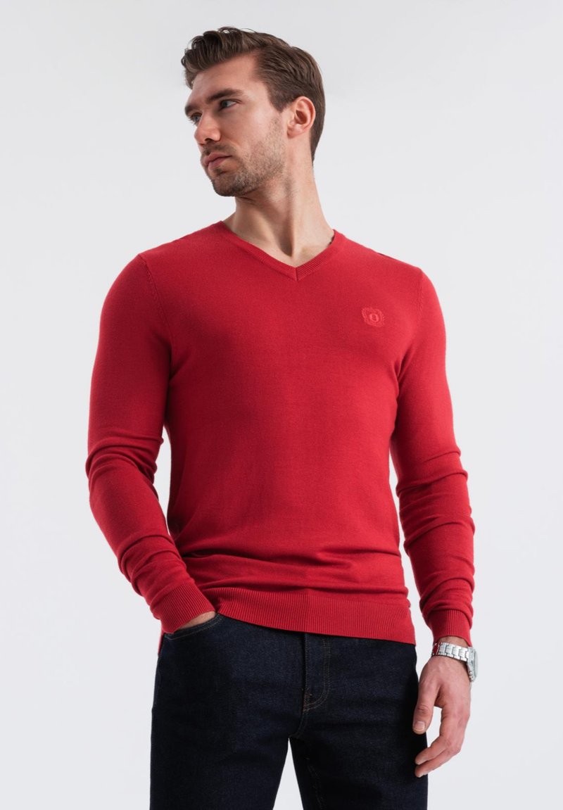 Maglione rosso a V in un morbido tessuto a maglia, con maniche lunghe e orlo a coste. Piccolo logo sul petto, abbinato a jeans scuri.