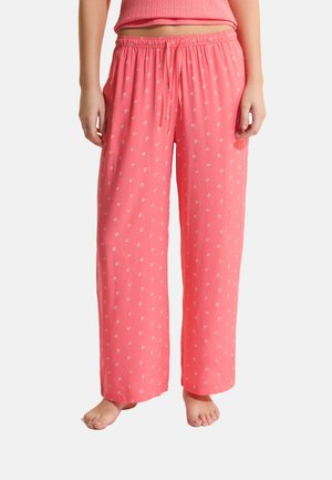 Pantaloni larghi rosa corallo con piccolo motivo floreale bianco, vita elasticizzata con coulisse, indossati da una persona in piedi scalza.