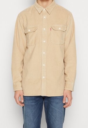 Chemise beige en velours côtelé à boutons avec deux poches poitrine portée avec un jean bleu, montrée des épaules aux hanches.