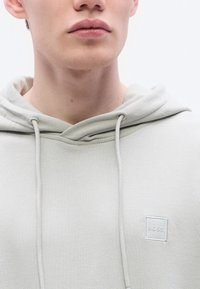 Ανοιχτό γκρι φούτερ με κουκούλα, κατασκευασμένο από απαλή ύφανση, με λογότυπο σε σχήμα τετραγώνου στο στήθος και κορδόνια σε αντίθετο χρώμα.