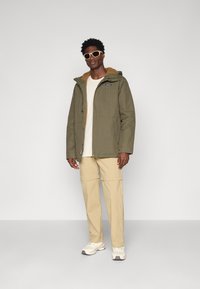 Olivenfarvet jakke med en struktureret sandfarvet foret, lynlås lukning og lommer, parret med beige bukser og hvide sneakers.