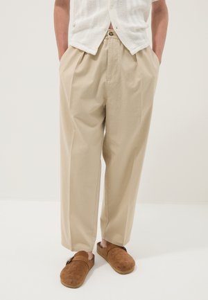 Hombre con pantalones beige holgados, camisa de punto blanca de manga corta y zapatos de ante marrón sin cordones, de pie con las manos en los bolsillos.
