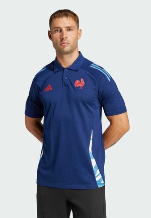 Polo de color azul marino en tejido transpirable con un logo de gallo rojo, acentos a rayas blancas y azules a los lados, y tres rayas azules en los hombros.