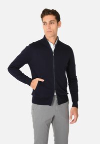 Cardigan zip-up blu navy con polsini e orlo a coste, caratterizzato da una trama liscia e due tasche frontali, indossato sopra una camicia con colletto.