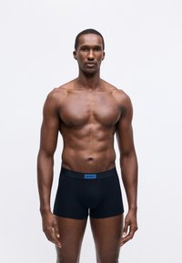 Muscular man standing facing forward wearing black boxer briefs with blue Hugo logo on plain white background.