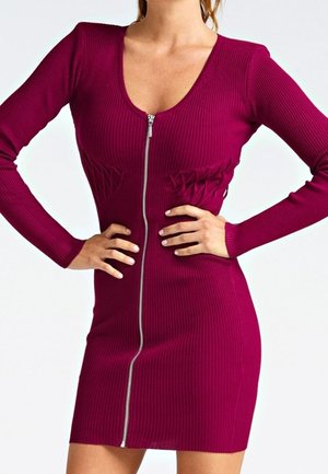 Kobieta ubrana w dopasowaną, prążkowaną, długorękawową bordową sukienkę z przednim zamkiem błyskawicznym i subtelnymi teksturowanymi detalami w talii, dłonie spoczywają na biodrach.