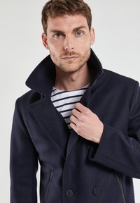 Cappotto doppiopetto in misto lana di colore blu navy con collo alto, due tasche frontali e dettagli in bottone nero, indossato sopra una camicia a righe.