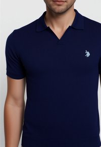 Polo de color azul marino hecho de una tela suave, con cuello clásico, mangas cortas y un logotipo bordado en azul claro en el pecho.