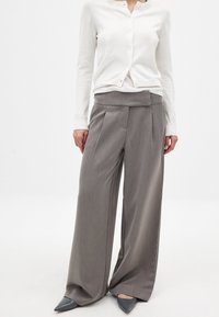 Pantalones grises de pierna ancha con cintura alta y frente plisado. Combinados con un cárdigan blanco de botones y zapatos grises de punta.