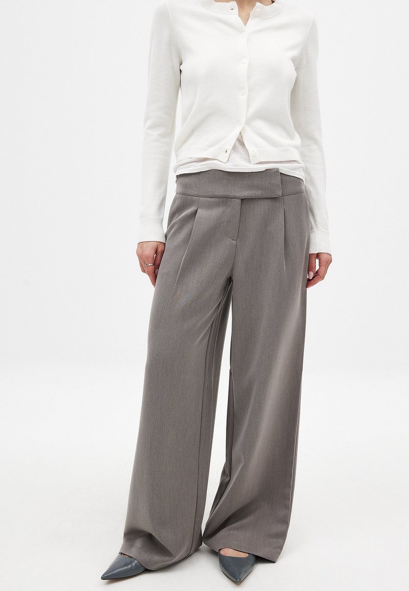 Pantalones grises de pierna ancha con cintura alta y frente plisado. Combinados con un cárdigan blanco de botones y zapatos grises de punta.