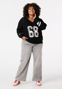 Glunderende vrouw met krullend haar in een zwarte gebreide trui met "68", grijze wijde jeans en roze sneakers, staand met één hand in haar zak.