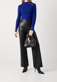Femme portant un haut à manches longues côtelé bleu avec des boutons, un pantalon large en cuir noir, des bottes noires pointues, tenant un petit sac à main noir.